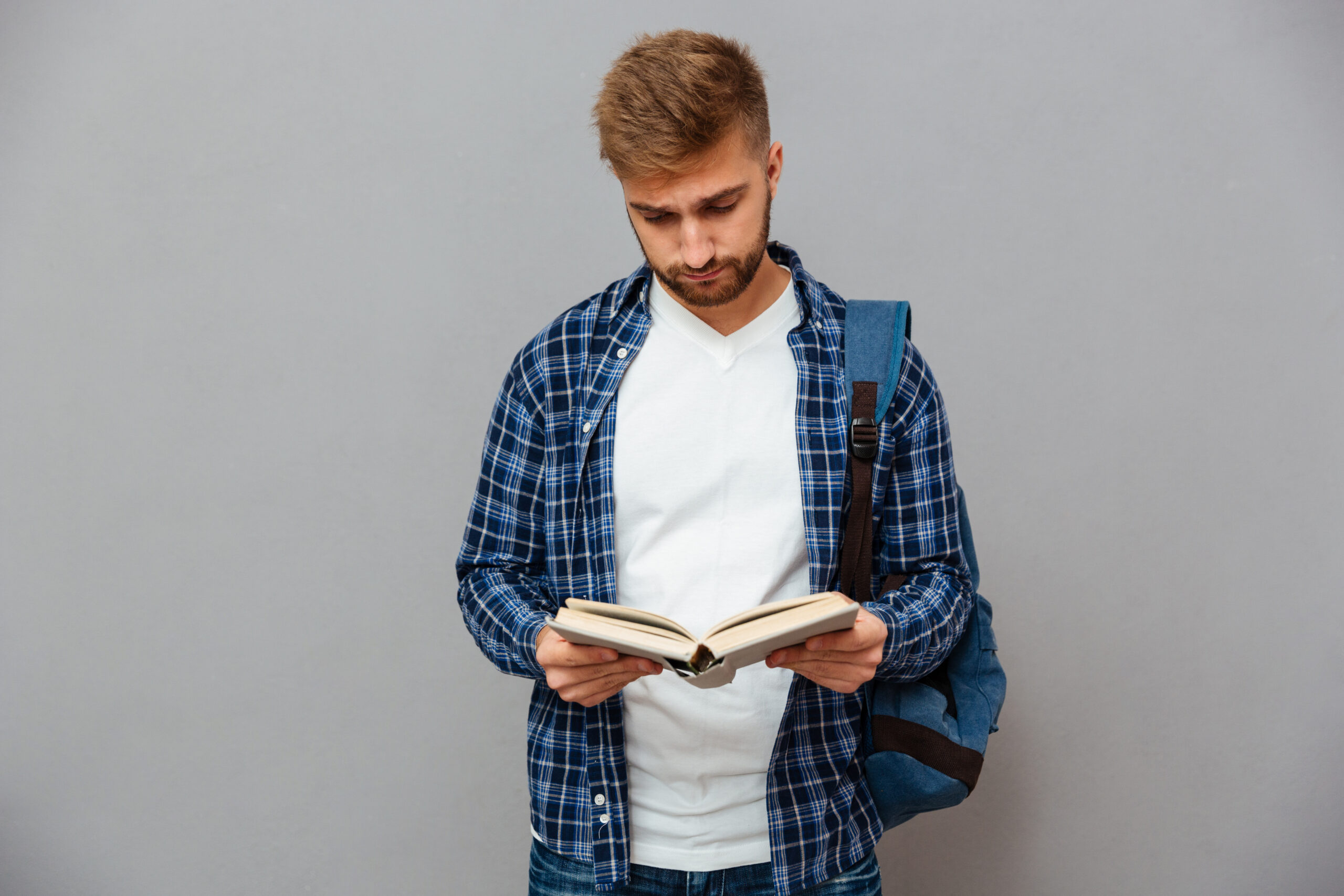 stylish-young-man-in-checkered-shirt-with-backpack-reading-book-on-gray-